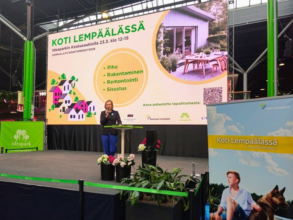 Kunnanjohtajja Heidi Rämö Ideaparkin Keskusaukion esintyymislavalla puhumassa. Taustalla keltasävyinen mainos Koti Lempäälässä tapahtumasta