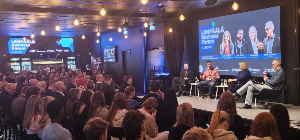 Suuri yleisö seuraa lavalla istuvien puhujien keskustelua modernissa tapahtumatilassa. Taustalla näkyy valaistu näyttö ja tapahtuman logo, tilassa on lämpimät valot ja mustat seinäpinnat.