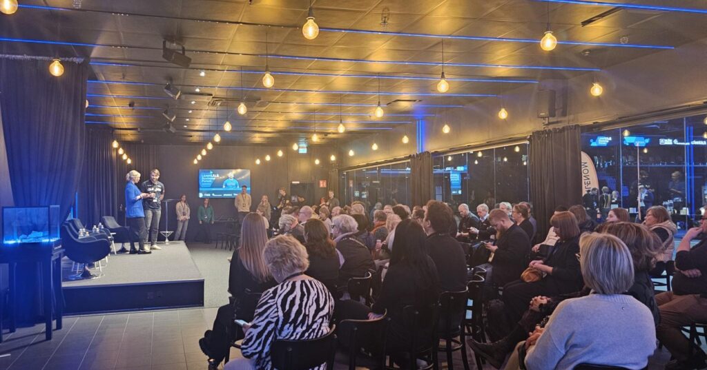 Tapahtumatila, jossa yleisö seuraa lavalla kahden henkilön keskustelua. Lava on valaistu lämpimillä kattovaloilla, taustalla näkyy näyttö ja tummat verhot, tilassa on moderni ja tunnelmallinen valaistus.